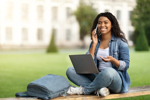 Улыбающаяся молодая чернокожая женщина разговаривает по мобильному телефону и пользуется ноутбуком на открытом воздухе