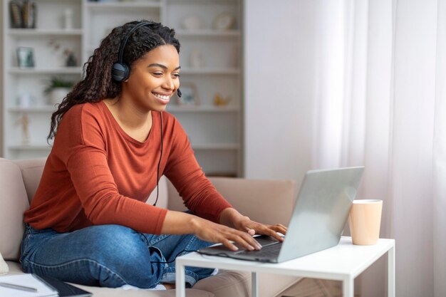 ヘッドセットをかぶった笑顔の若い黒人女性が自宅でラップトップコンピュータを使用しています