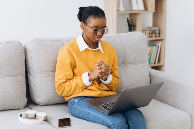 Smiling young black woman in yellow sweater freelancer working from home, sitting on sofa, using laptop, copy space and thinking seriously.