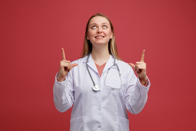 Photo smiling young blonde female doctor wearing medical robe and stethoscope around neck looking and pointing up