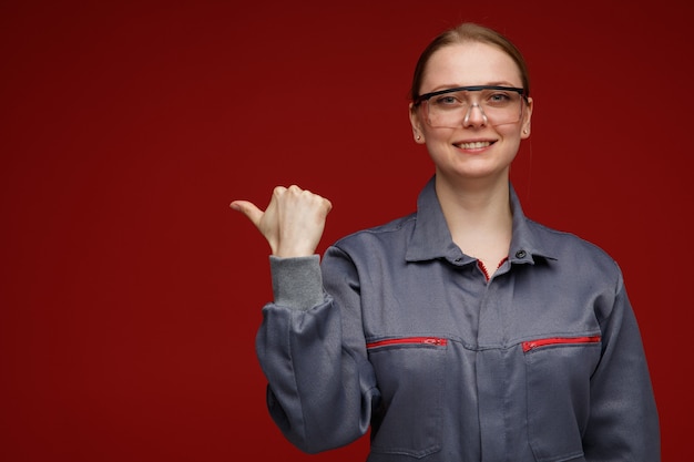 Smiling young blonde female engineer wearing uniform and safety glasses pointing to side 