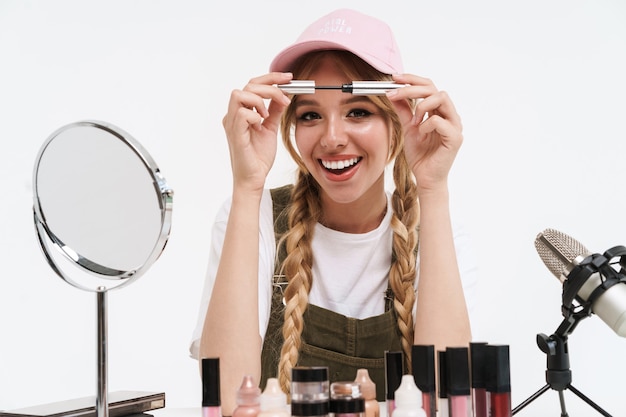 Sorridente giovane ragazza bionda blogger che fa un video di bellezza con il trucco mentre è seduto al tavolo isolato su bianco