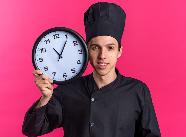 Smiling young blonde male cook in chef uniform and cap holding clock touching face with it 