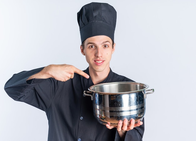 Sorridente giovane cuoco maschio biondo in uniforme da chef e berretto che tiene e punta alla pentola