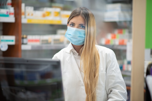 Smiling young blonde pharmacist wearing covid coronavirus mask inside her pharmacy