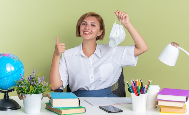 Smiling young blonde student girl sitting at desk with school tools holding protective mask looking at camera showing thumb up isolated on olive green wall