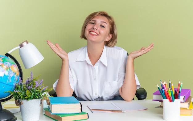 Smiling young blonde student girl sitting at desk with school tools looking at camera showing empty hands isolated on olive green wall