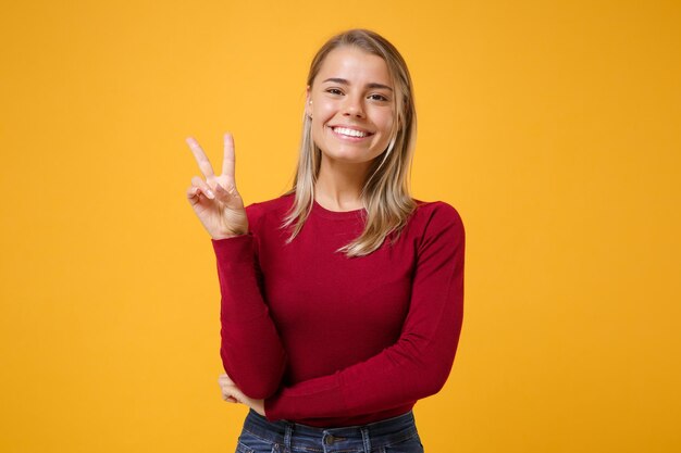 Smiling young blonde woman girl in casual clothes posing isolated on yellow orange wall background studio portrait. People sincere emotions lifestyle concept. Mock up copy space. Showing victory sign.