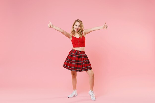 Smiling young blonde woman girl in red sexy clothes posing isolated on pastel pink wall background studio portrait. People sincere emotions lifestyle concept. Mock up copy space. Showing thumbs up.