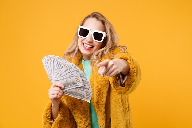 Smiling young blonde woman girl in yellow fur coat, dark sunglasses isolated on orange background. people lifestyle concept. mock up copy space. hold fan of cash money pointing index finger on camera