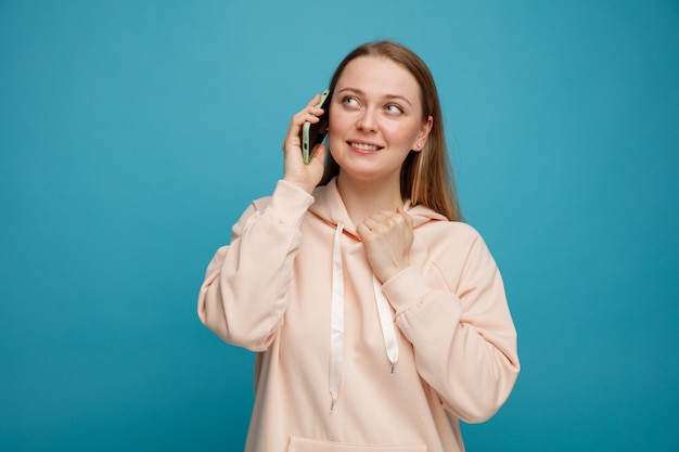 Smiling young blonde woman looking up at corner grabbing collar of her sweatshirt talking on phone 