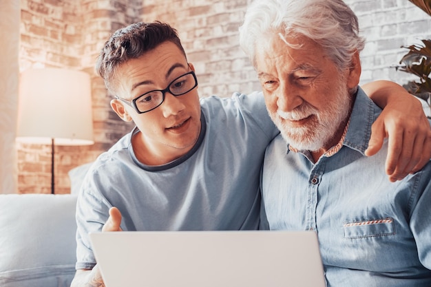 Smiling young boy helping senior grandfather browse with laptop
and choose goods or services on web surfing together at home
younger generation caring about older relatives teaching using
computer