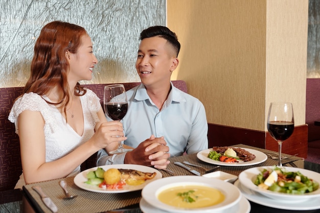 Smiling young boyfriend and girlfriend in love holding hands when eating dinner and talking at restaurant table