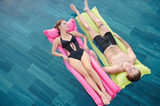 Smiling young boyfriend and girlfriend relaxing on floating mattresses in swimming pool