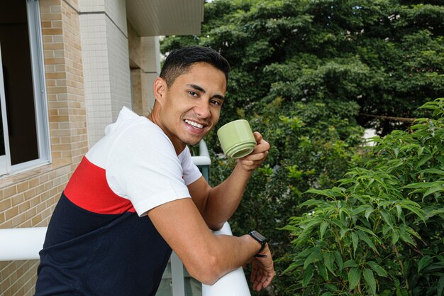 建物のバルコニーの手すりに寄りかかって、コーヒーを飲みながら笑顔の若いブラジル人。