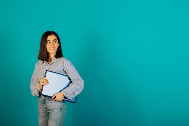 Smiling young brunette girl in blue shirt and jeans are posing isolated on blue surface.