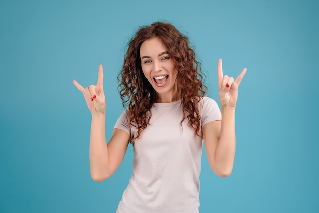 Smiling young brunette girl isolated over blue background showing rock gestures with her hands