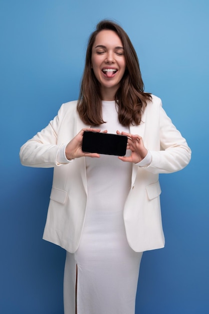 Smiling young brunette with long hair woman in white dress showing smartphone screen with mockup