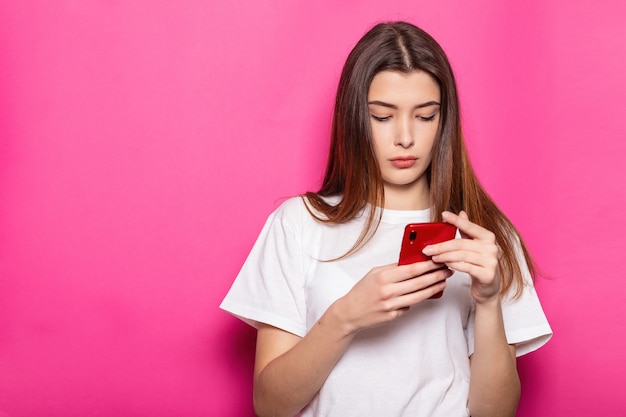 Smiling young brunette woman girl posing isolated on pastel pink wall background studio portait. People lifestyle concept. Mock up copy space. Using mobile phone, typing sms message