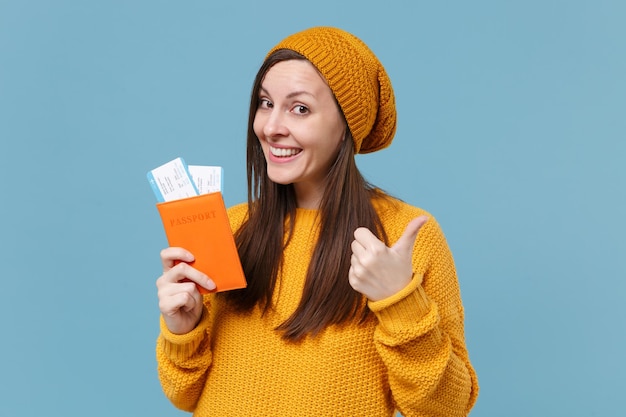 Smiling young brunette woman girl in sweater hat posing isolated on blue background studio portrait. People lifestyle concept. Mock up copy space. Hold passport boarding pass tickets showing thumb up.