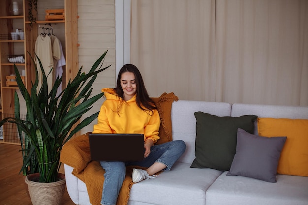 A smiling young brunette woman with long hair in a yellow\
sweatshirt, blue jeans and white sneakers sits at home on a gray\
sofa with a laptop. there is a mustard blanket on the sofa. home\
green plants