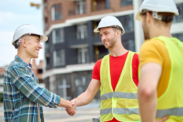 Smiling young builder and his coworker dressed in protective helmets and safety vests greeting a middle-aged male engineer