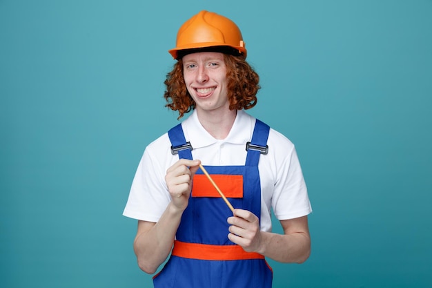 Smiling young builder man in uniform holding pencil isolated on blue background