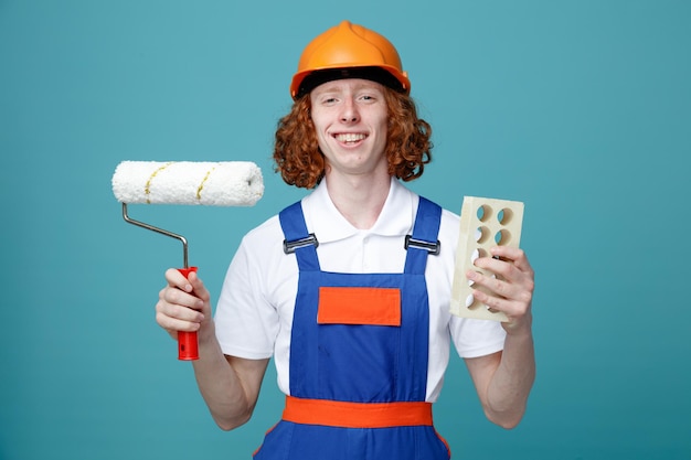 Smiling young builder man in uniform holding roller brush and brick isolated on blue background