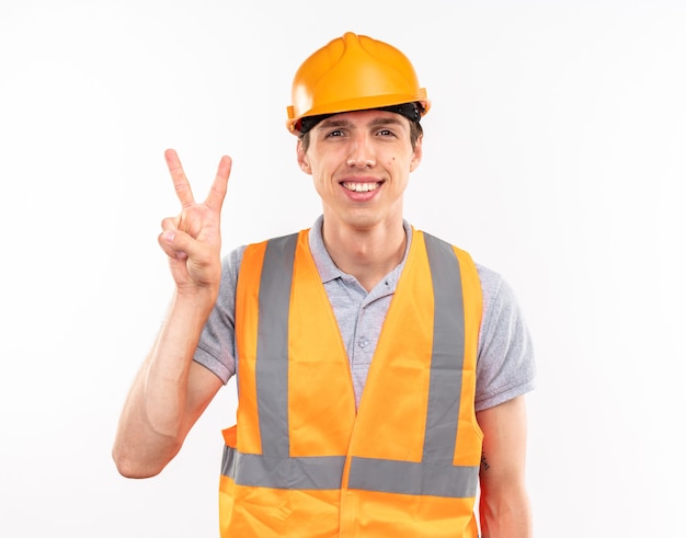 Smiling young builder man in uniform showing peace gesture isolated on white wall