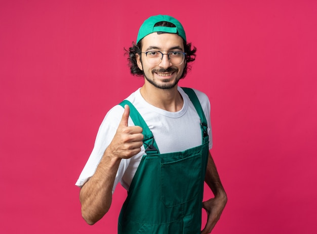 Smiling young builder man wearing uniform with cap showing thumb up 