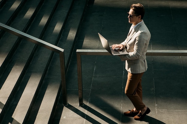 Улыбающийся молодой деловой человек проводит время на свежем воздухе в городе, используя ноутбук на лестнице
