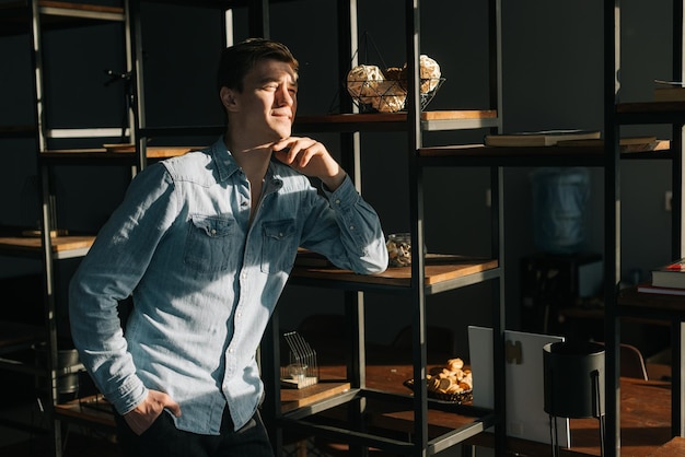 Smiling young business man wearing fashion casual clothing standing near shelves in modern office room looking away bright sunlight