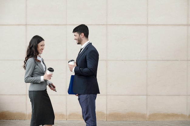 Smiling young business people standing outdoors drinking take\
out coffee and discussing news and gos...