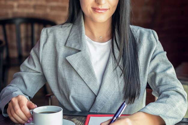 Smiling young business woman is writing to notebook drinking coffee in cafe. Business or work concept.