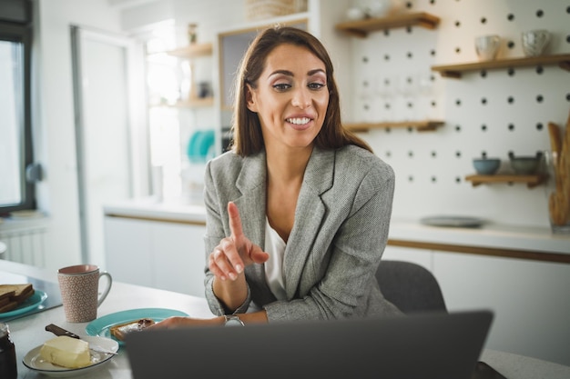 Smiling young business woman making video call and heaving a breakfast in her kitchen.