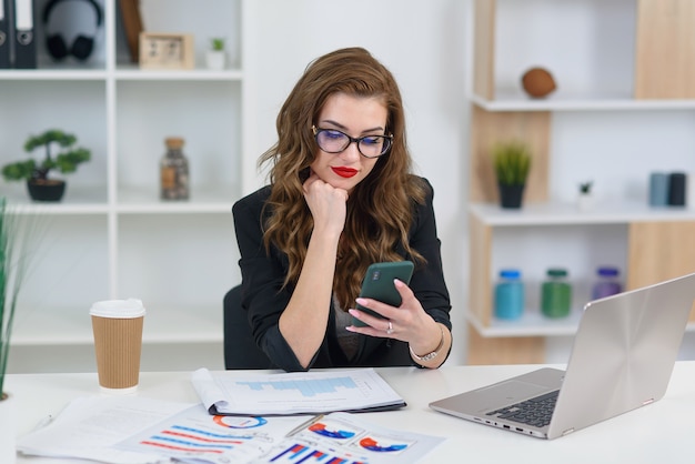 Smiling young business woman in stylish clothes sitting in cozy modern office and browsing something on smartphone.