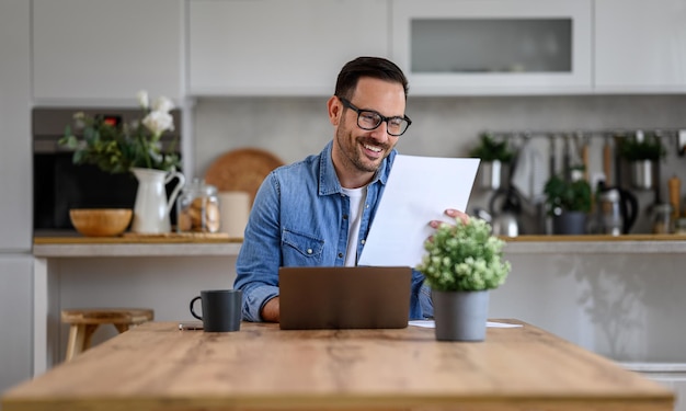 Smiling young businessman analyzing report and working over laptop on desk in home office