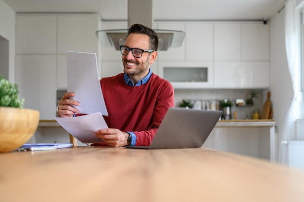 Smiling young businessman reading document while sitting with laptop at desk in home office