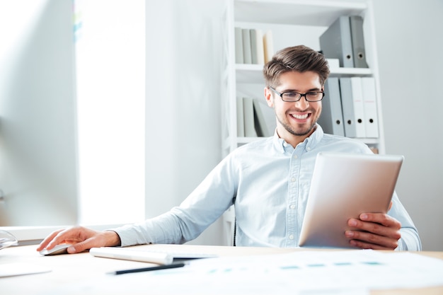 Sorridente giovane uomo d'affari seduto al tavolo e utilizzando il computer tablet in ufficio