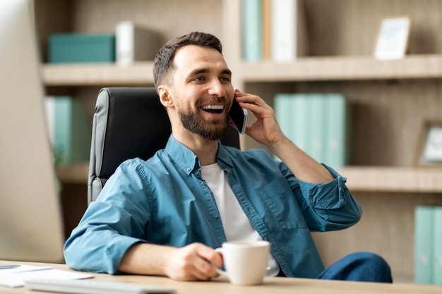 Smiling young businessman talking on cellphone and drinking coffee at workplace