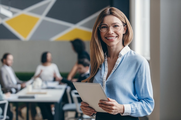 Smiling young businesswoman holding digital tablet while her colleagues working on background