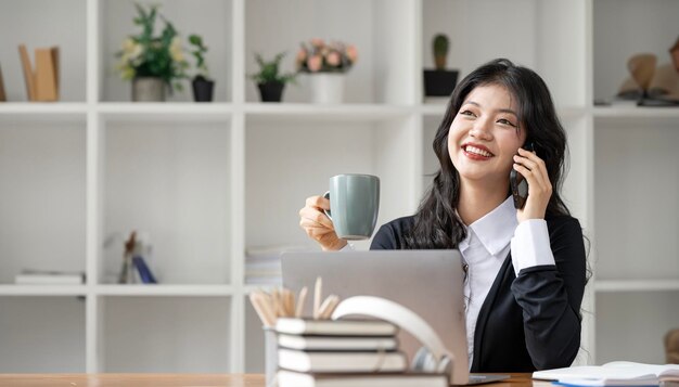 Smiling young businesswoman holding mug and using smart phone sitting at home office