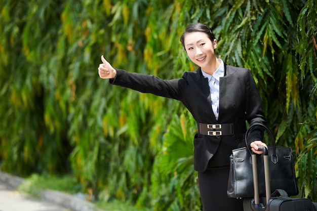 Smiling young businesswoman making thumbs-up gesture when trying to catch a taxi car to airport or hotel