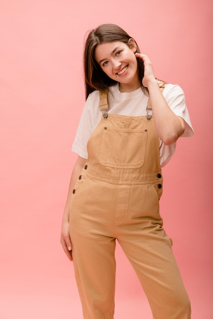 Smiling young caucasian brunette girl in tshirt and overalls looks at camera on pink background Lifestyle female beauty concept