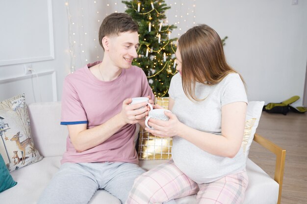 Smiling young Caucasian couple holding a coffee cup at home in Christmas eve holiday with Christmas tree background.