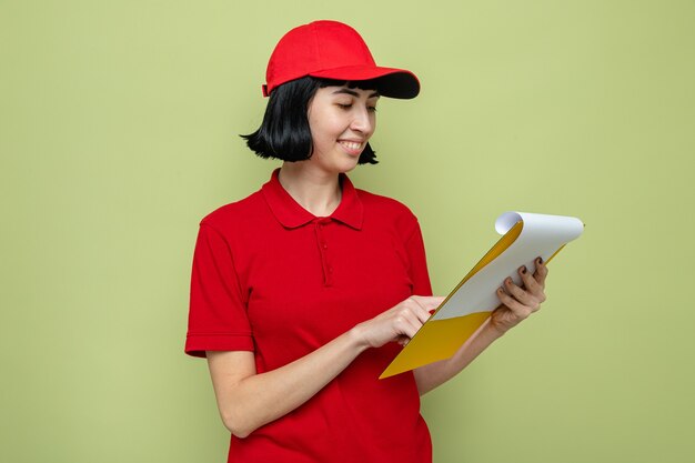 Smiling young caucasian delivery girl holding and looking at clipboard