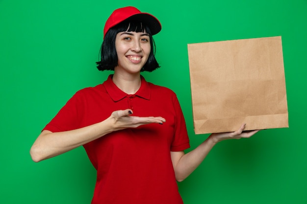 Улыбающаяся молодая кавказская курьерская девушка держит и указывает рукой на бумажную упаковку пищевых продуктов