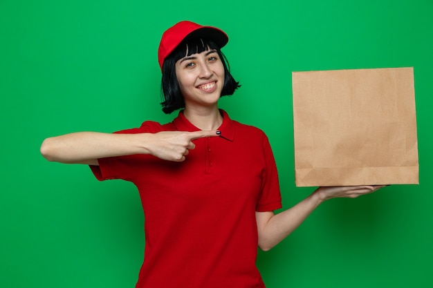 Smiling young caucasian delivery girl holding and pointing at paper food packaging