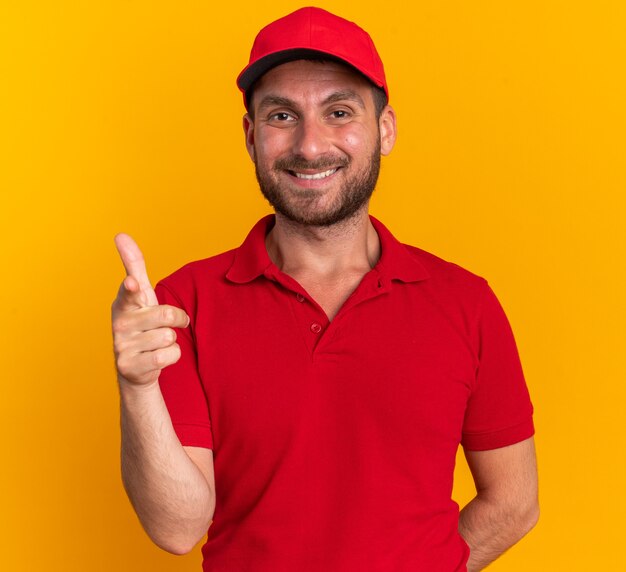 Smiling young caucasian delivery man in red uniform and cap keeping hand behind back looking and pointing at camera isolated on orange wall