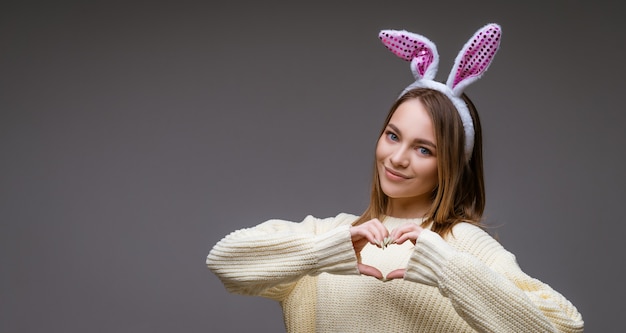 Улыбающаяся молодая кавказская девушка, блондинка с кроличьими ушками, показывает сердце двумя руками и смотрит в камеру, изолированную на сером фоне